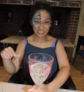 Myah, a guest with a learning disability, enjoying a supported holiday at Nutley Edge. She has a sparkly painted face and is holding a drink in a flamingo glass. 